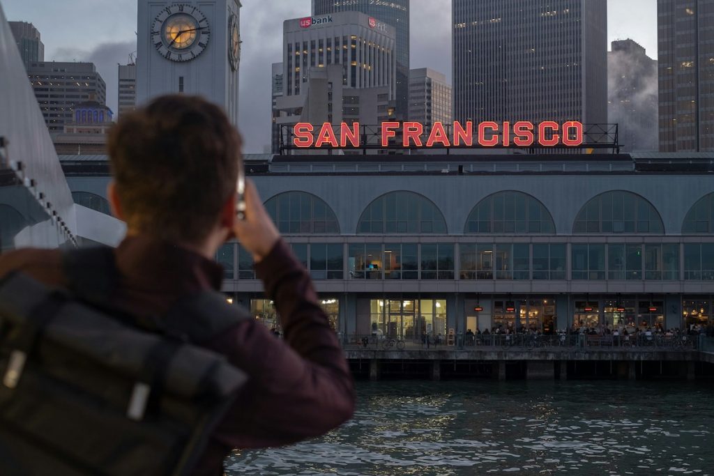 A person with a backpack takes a picture of the glowing San Francisco sign on a waterfront building at dusk.