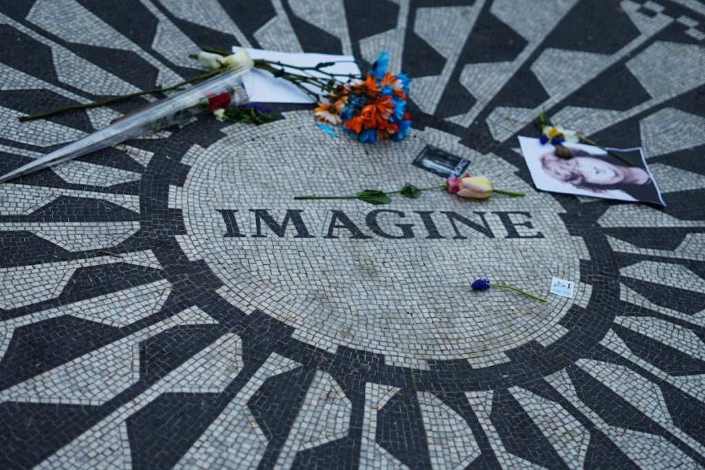 A geometric sunburst mosaic with “IMAGINE” at the center in Strawberry Fields, Central Park, is covered in fresh flowers and a tribute photograph.