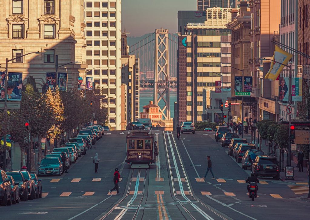 A classic San Francisco cable car climbs a steep hill as pedestrians cross, with the Bay Bridge and city buildings behind.