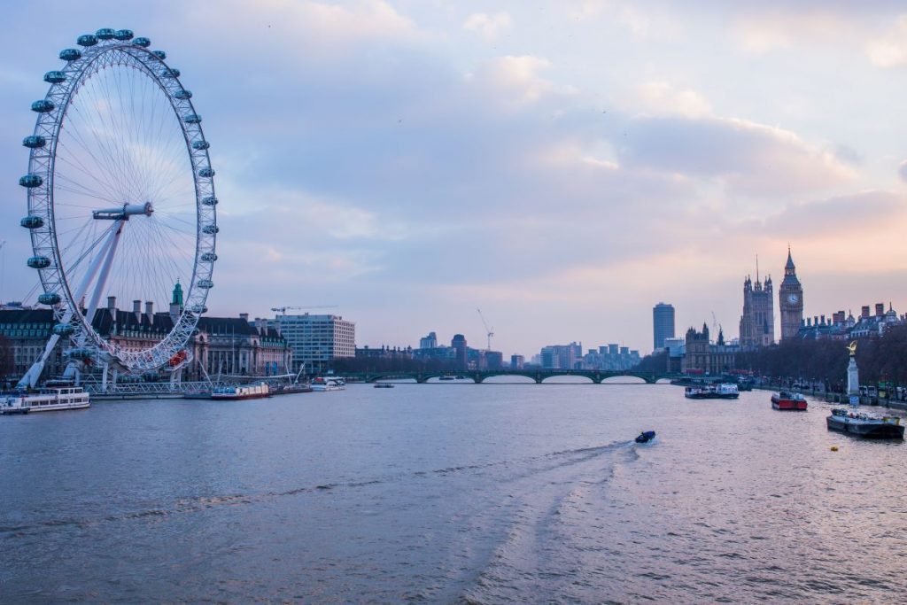 The London Eye stands beside the River Thames, boats travel below, and Big Ben and Parliament appear in the distance.