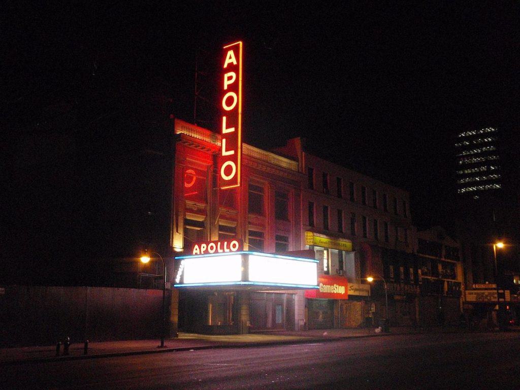 The Apollo Theater in Harlem glows at night, its bright red neon sign and marquee lighting up the dark street.