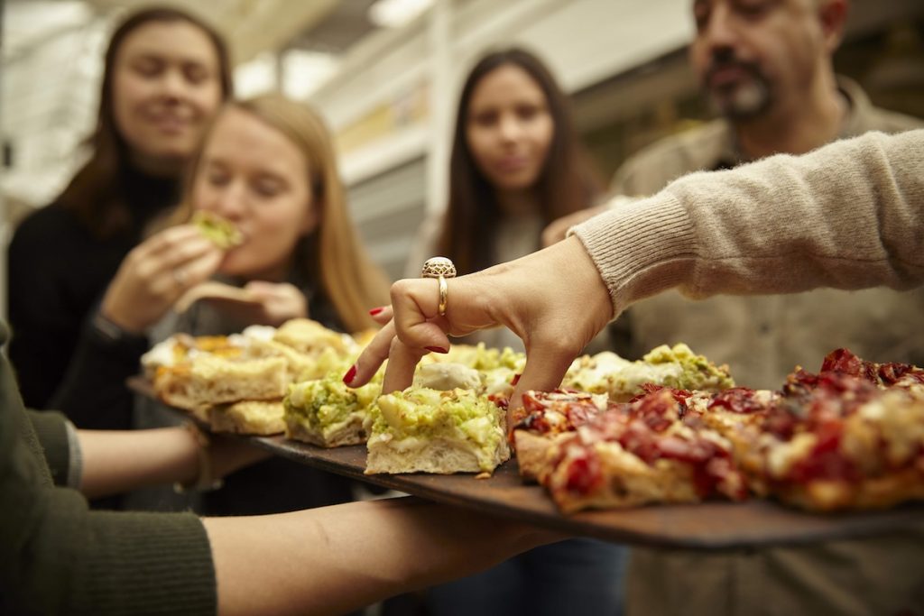 A hand reaches for a pizza slice on a tray while four friends gather around, two already enjoying their food at a table.