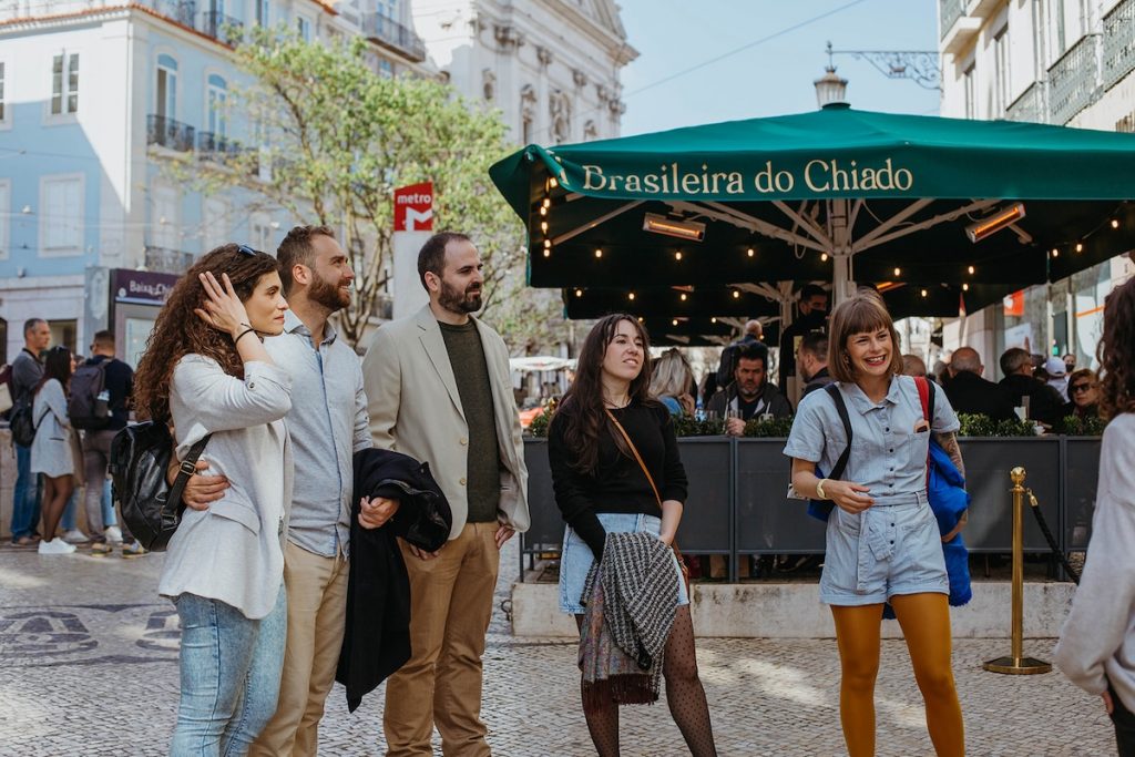 Five friends smile together in Lisbon’s bustling Chiado plaza near Café A Brasileira, with classic buildings and a metro sign behind them.