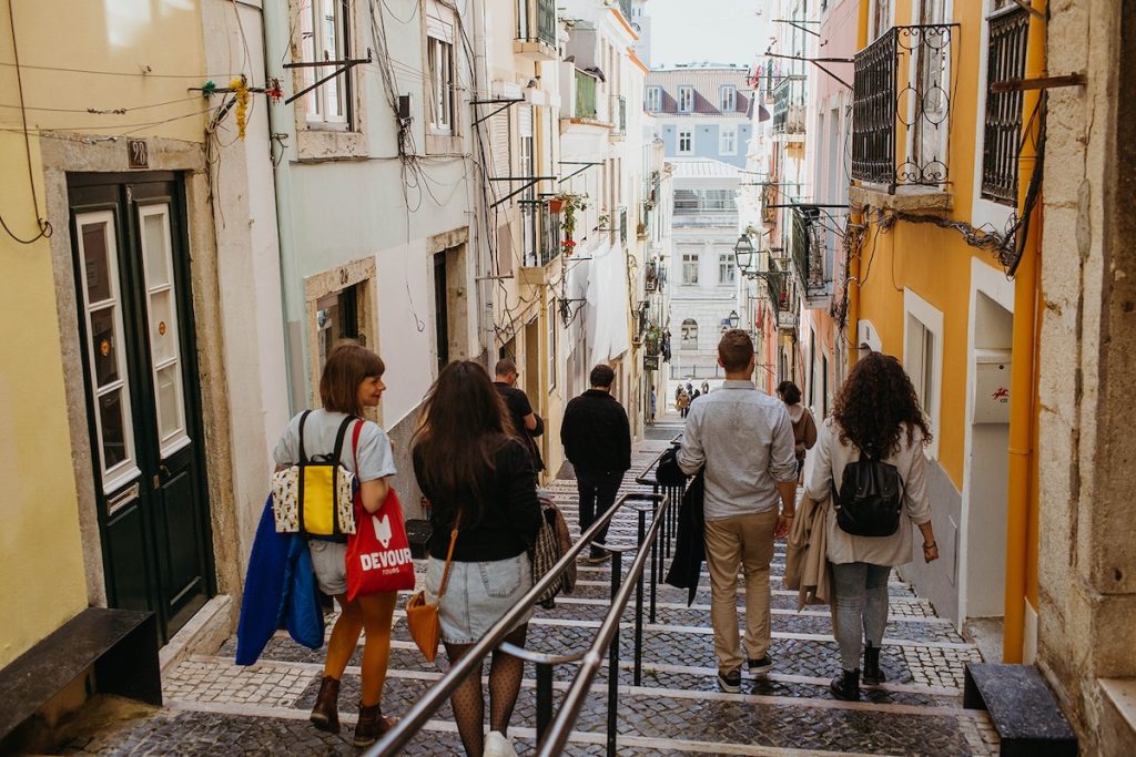Several people stroll along a narrow cobblestone street with colorful buildings and balconies, approaching a sunlit plaza in Old Havana.