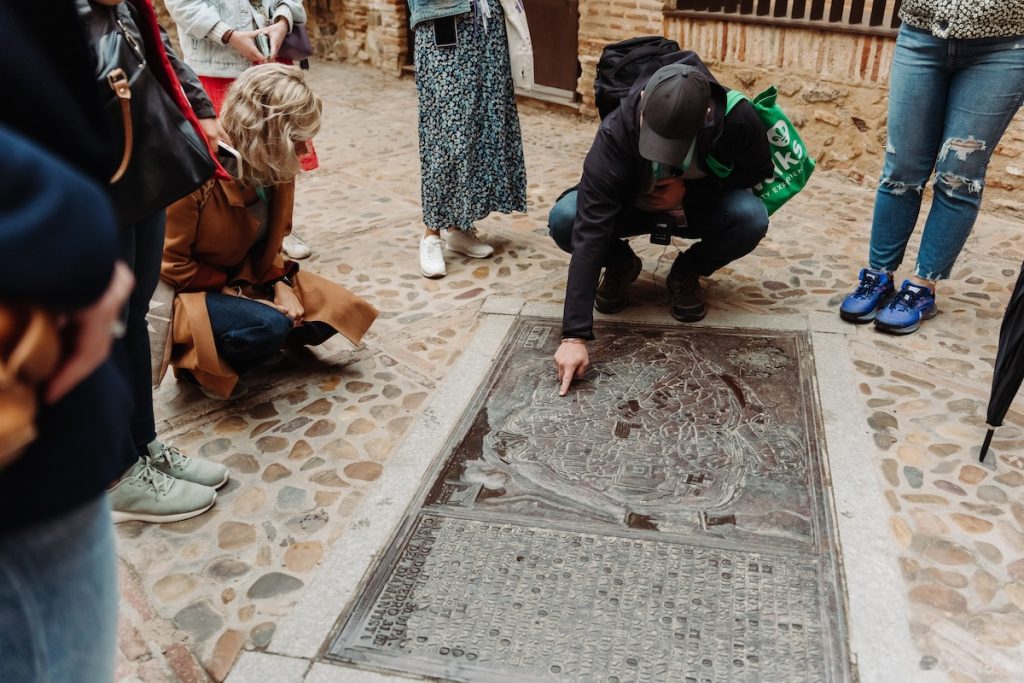 A guide kneels and points at a detailed bronze relief map embedded in cobblestone as tourists listen attentively during a city tour.