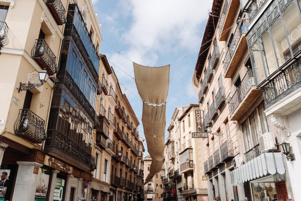 A narrow street in a historic European city has colorful fabric canopies overhead, with balconies, shops, and partly cloudy skies above.