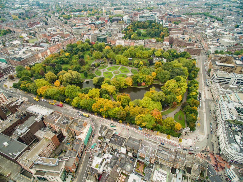 Aerial view of Central Park in New York City, showing green trees, winding paths, ponds, and surrounding skyscrapers under clear skies.