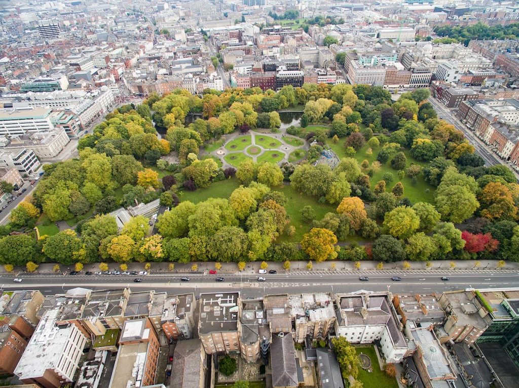 Aerial view shows a circular city park with winding paths and dense trees, surrounded by busy streets and tall buildings.