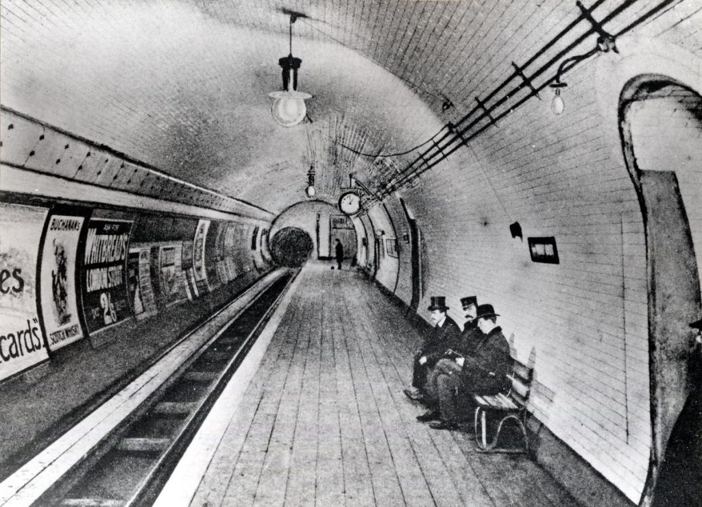 Three men in hats sit quietly on a bench at a nearly empty London Underground platform, surrounded by vintage posters and arched lights.