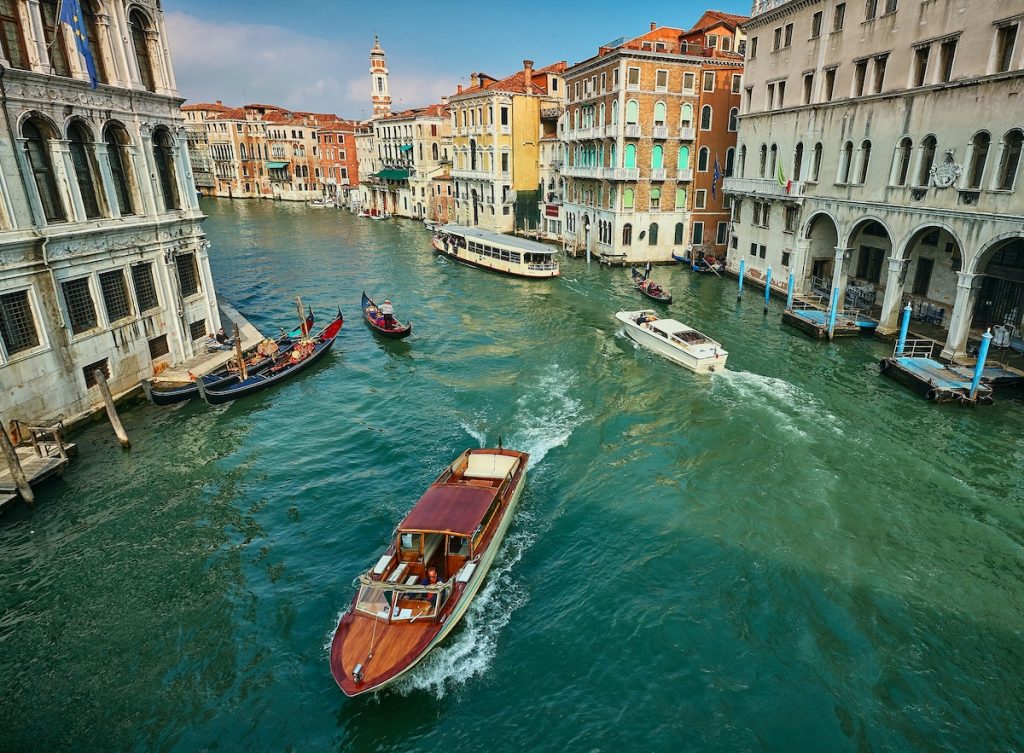 Boats and gondolas move on Venice’s Grand Canal, passing historic, colorful buildings beneath a bright blue sky with clouds.
