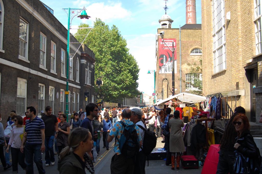 Crowds browse market stalls along a sunny London street, surrounded by brick buildings and greenery in the background.