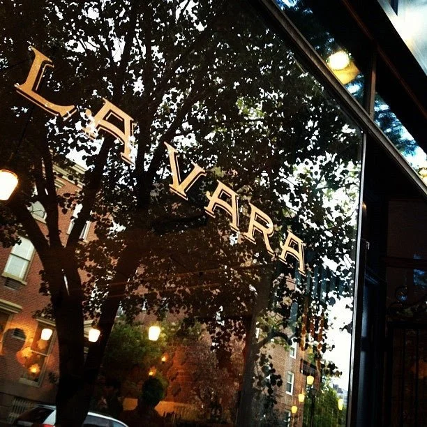 The glass window reading LA VARA reflects trees, streetlamps, and brick buildings at dusk on a Brooklyn food tour.