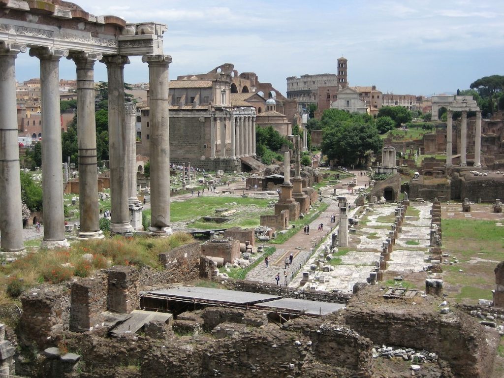 Tall columns and stone remains of the Roman Forum stand among greenery, with modern Rome visible under a partly cloudy sky.