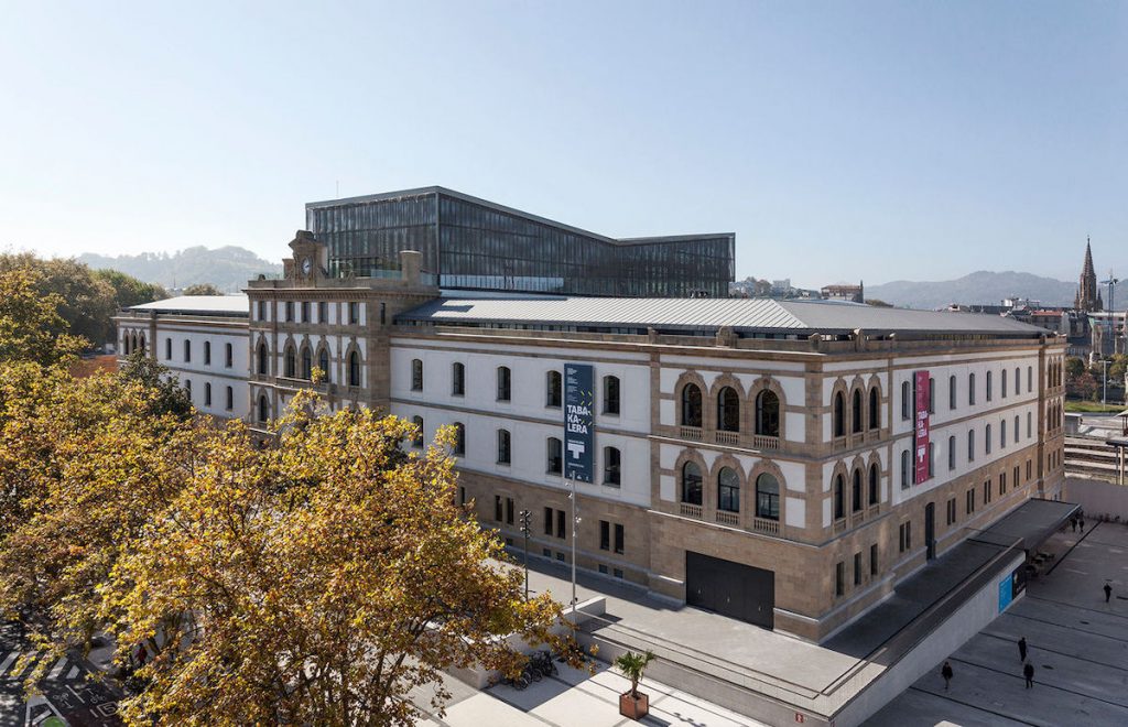 A grand historic building with modern glass rooftop additions stands in San Sebastián, Spain, surrounded by trees and pedestrians.