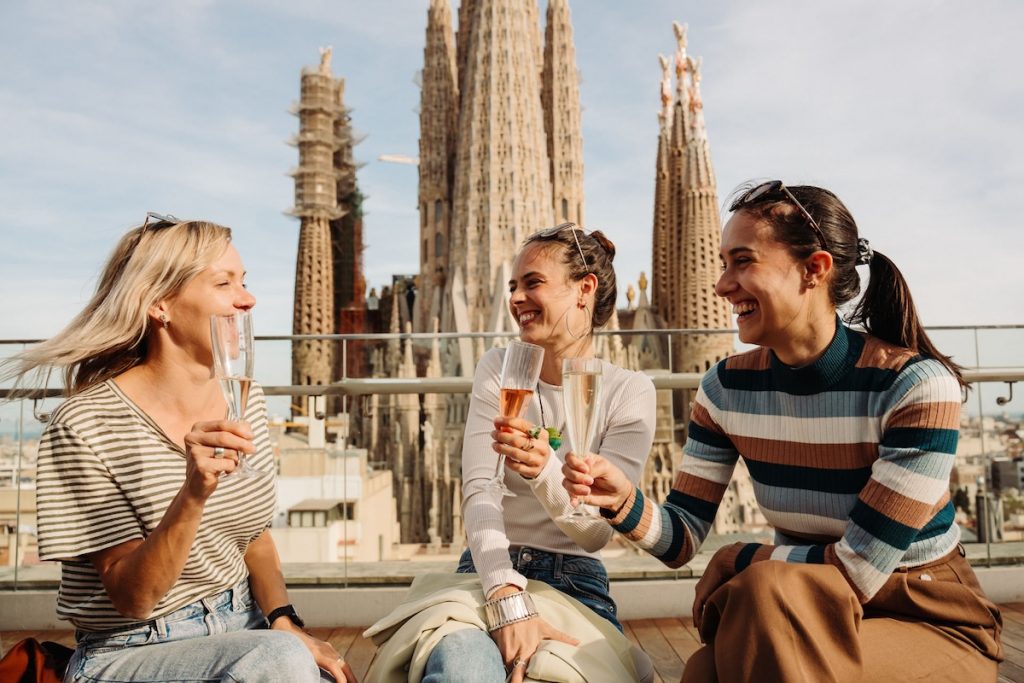 Three women toast with champagne, smiling together on a rooftop terrace, with Barcelona’s Sagrada Familia basilica rising in the background.