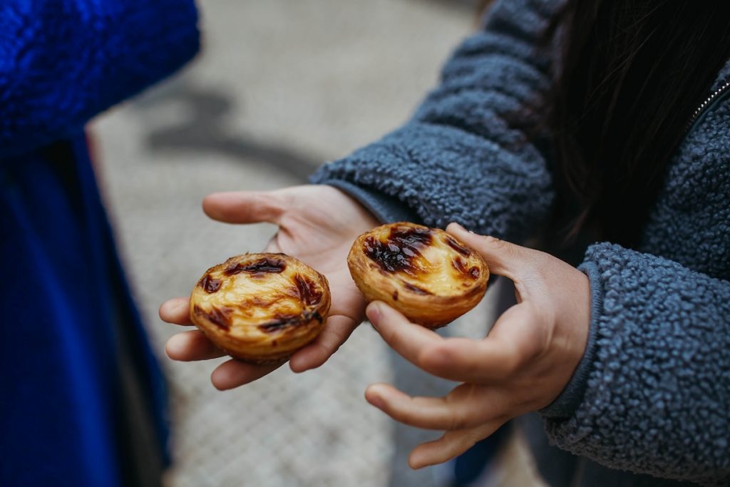 A person in a gray jacket holds two golden, caramelized pastéis de nata in their hands on a Lisbon street.