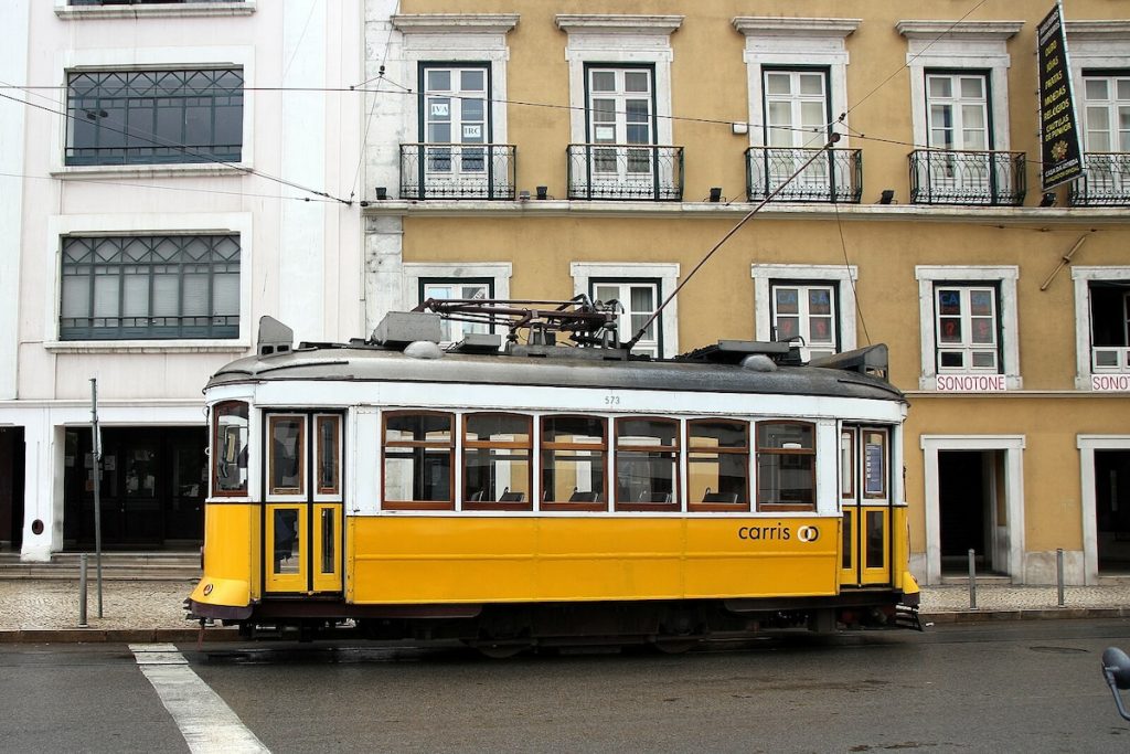A classic yellow Carris tram travels through Lisbon, Portugal, passing historic beige and white buildings with ornate balconies and large windows.