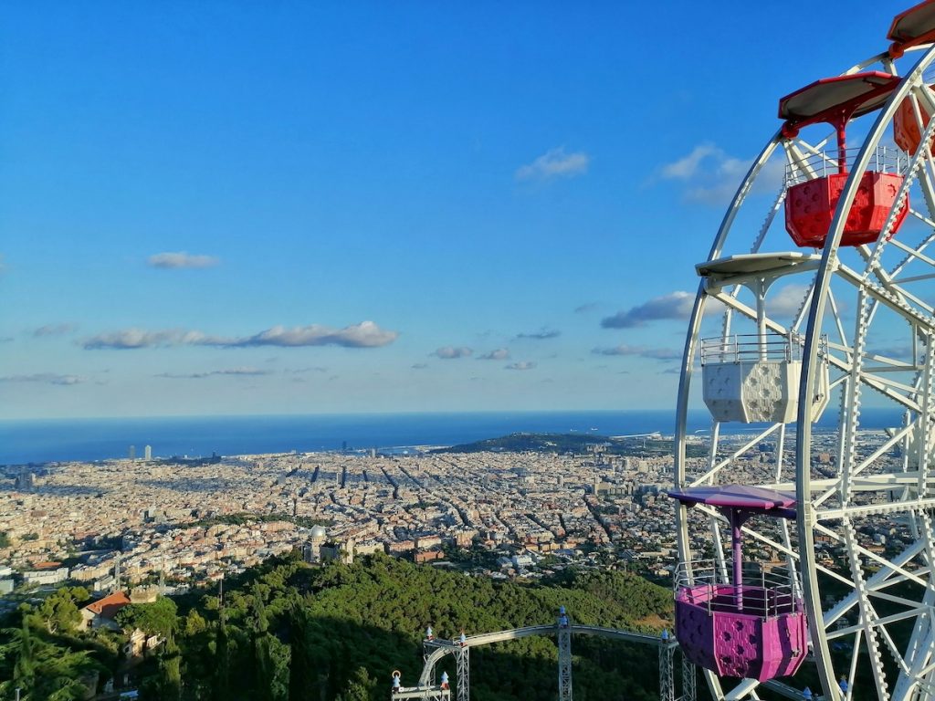 A red, yellow, and blue Ferris wheel overlooks Barcelona’s cityscape, lush green hills, and the Mediterranean Sea on a sunny day.