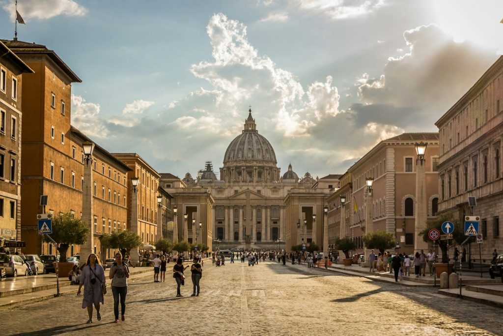 People walk on a cobblestone street toward St. Peter’s Basilica in Vatican City under dramatic sunlight breaking through clouds.