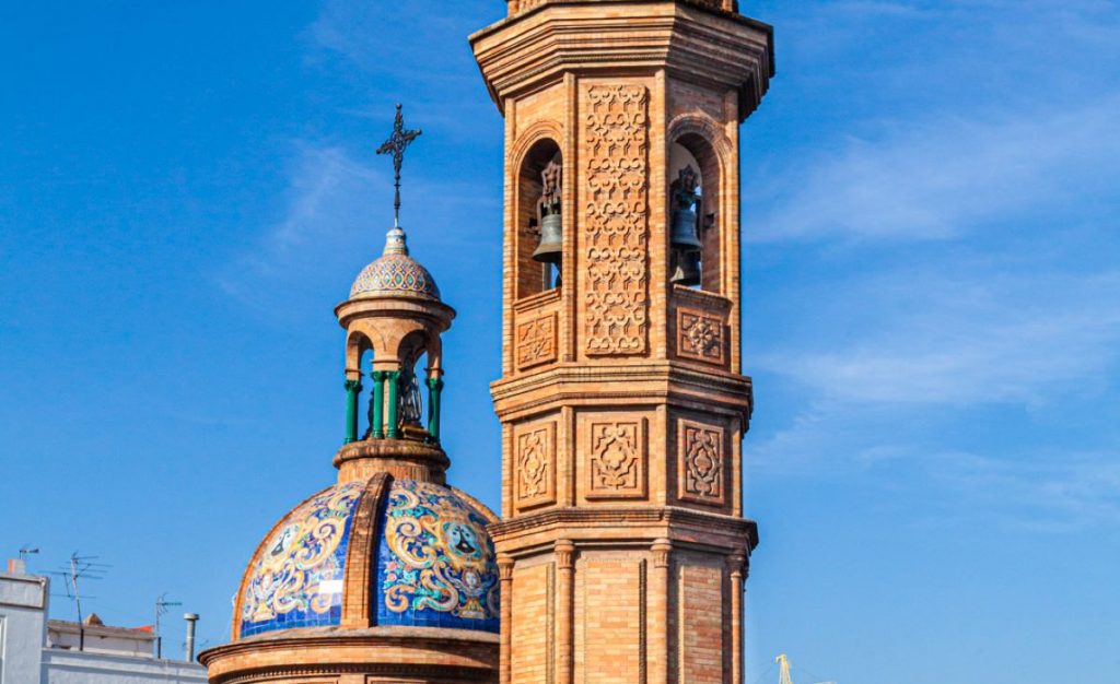 A woman cycles past the Church of El Salvador’s brick bell tower and blue-tiled dome under Seville’s clear sky.