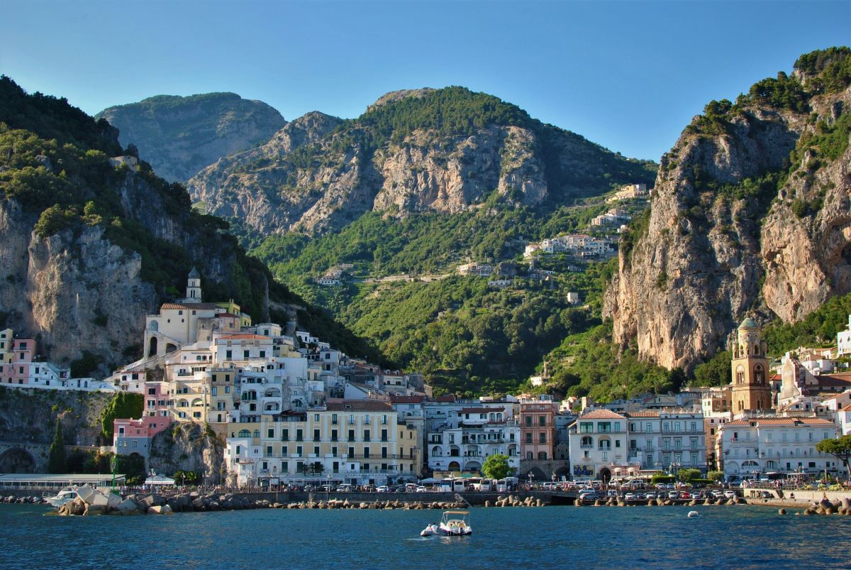 Colorful buildings cluster along Amalfi’s coastline beneath steep, green mountains as a small boat floats on the bright blue sea.