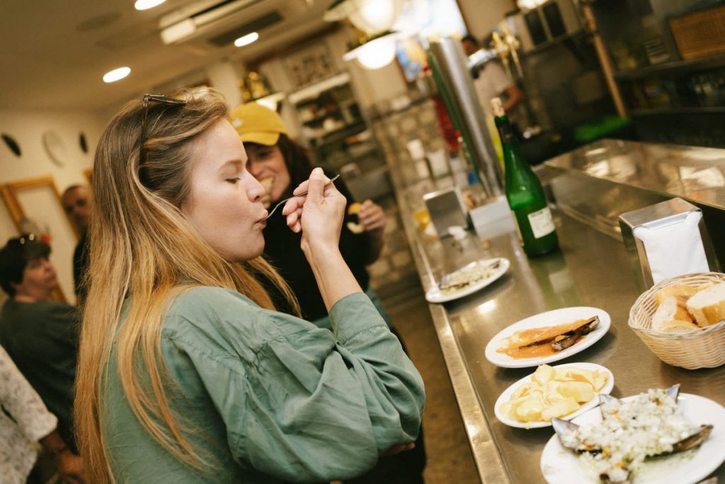 A woman with long blonde hair eats pintxos at a bustling San Sebastian bar, savoring food and cider on the counter.