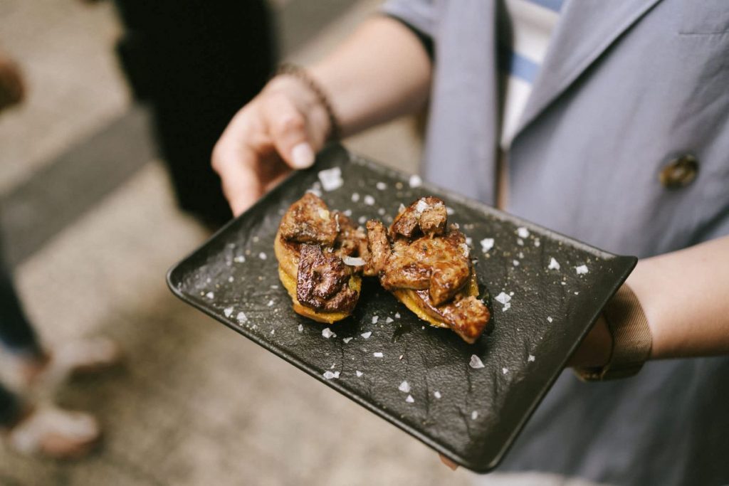 A person presents two pieces of grilled meat on toasted bread, topped with sea salt, in a setting reminiscent of San Sebastian pintxos.