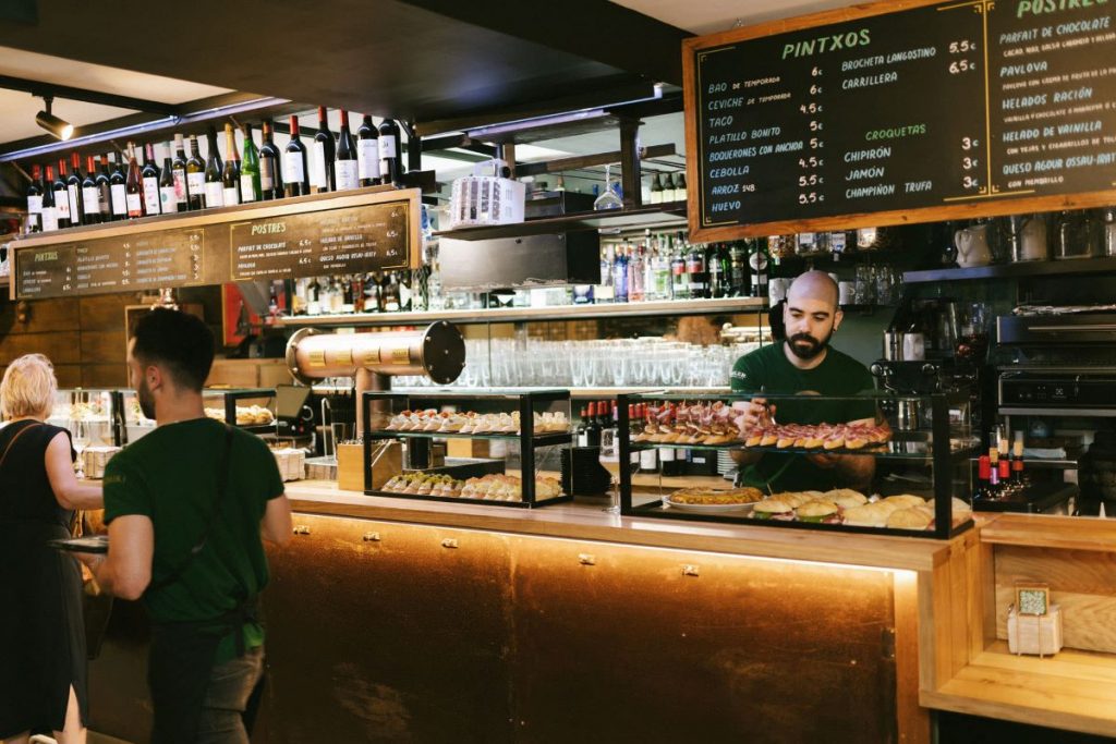 Two staff prepare food behind a tapas bar in San Sebastian, serving pintxos and pastries as a customer orders.