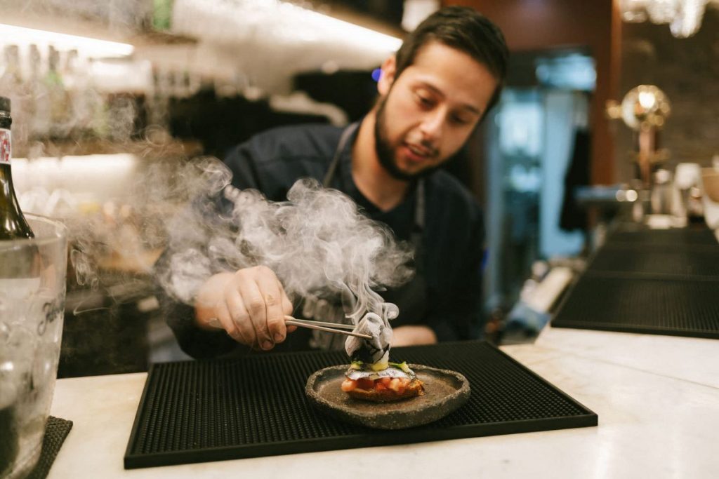 A chef in a modern San Sebastian restaurant uses tongs to garnish a smoky pintxo, showcasing Basque culinary artistry.