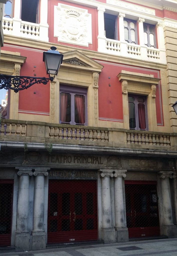 A red-walled historic building in San Sebastian Old Town features ornate stonework, three arched doors with grills, and a street lamp.