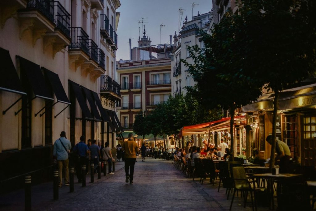 People walk and dine at outdoor cafes along a narrow street in Seville at dusk, with warm lights creating a lively atmosphere. A must do for any travel guide to Seville