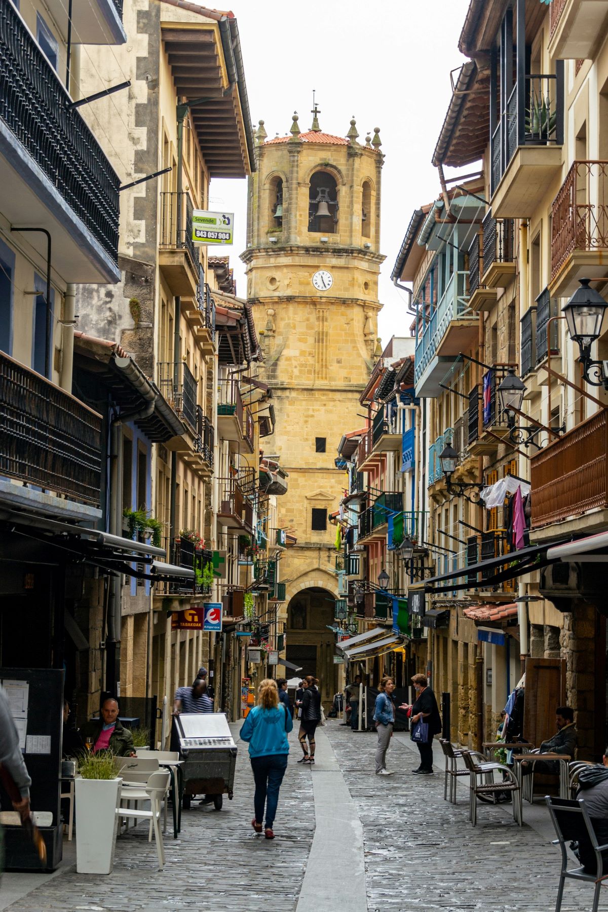 A group of people walking down a narrow street in Getaria, Spain near a church tower. 