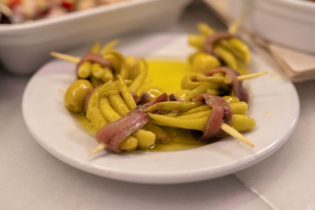 A white plate holds San Sebastian pintxos: green chili peppers, olives, and anchovies on skewers with olive oil; napkins behind.