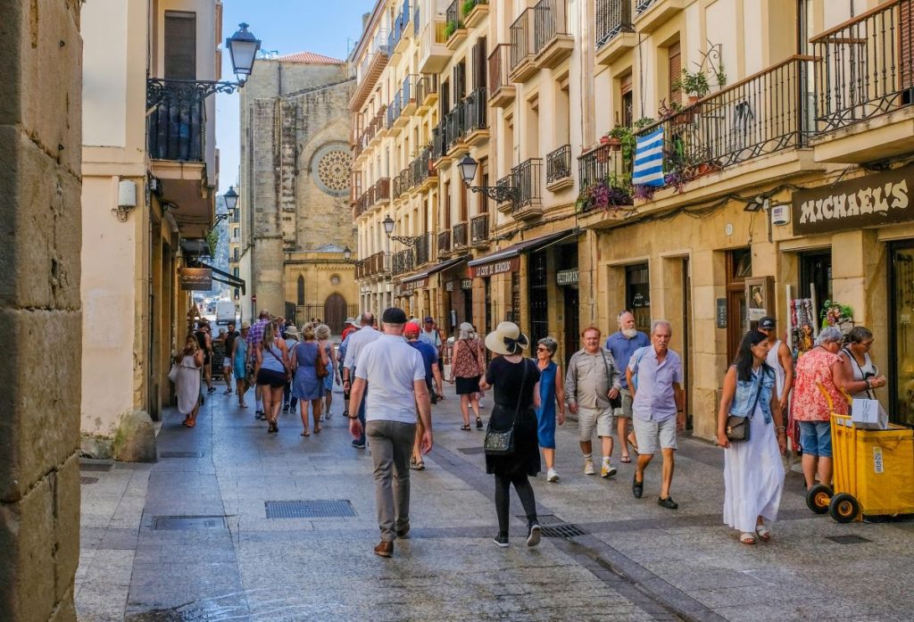 Shoppers stroll a lively, sunlit street with cafes and plant-filled balconies, leading towards the historic Santa Maria del Mar church in Barcelona.