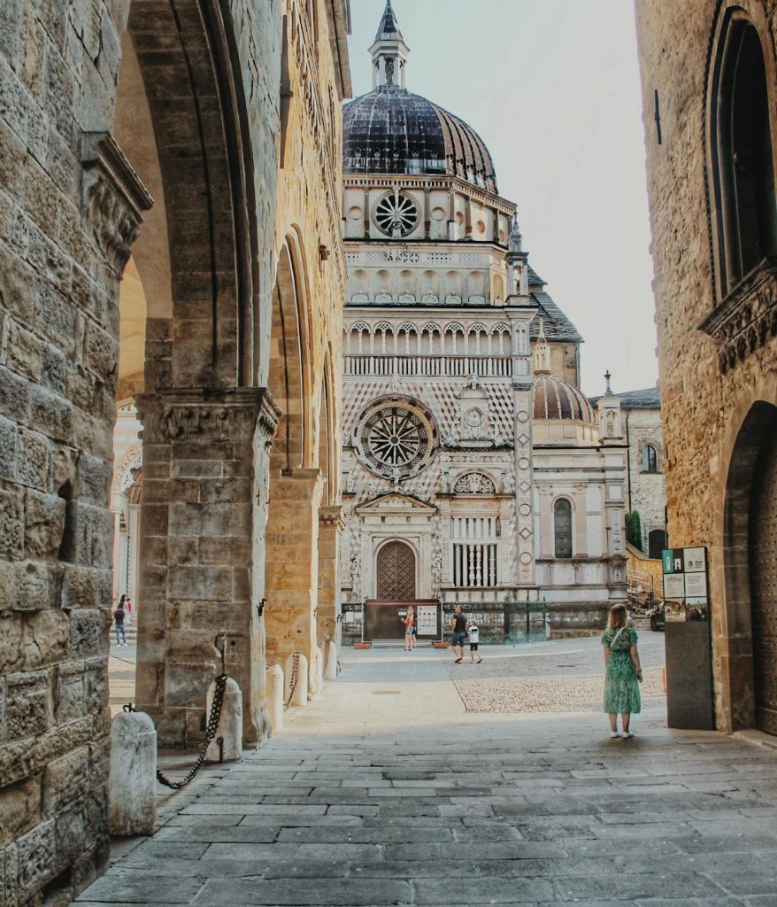 A woman in a green dress stands before Bergamo Cathedral’s marble facade and dome, admiring the architecture on a sunny day.