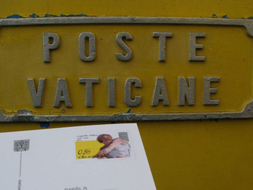 A hand holds an envelope with a Vatican stamp near a yellow metal mailbox marked POSTE VATICANE in Vatican City.