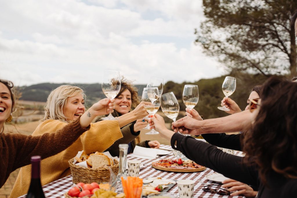 Four friends toast with wine at an outdoor table in Montserrat, Spain, sharing bread, fruit, and vegetables amid lush hills.