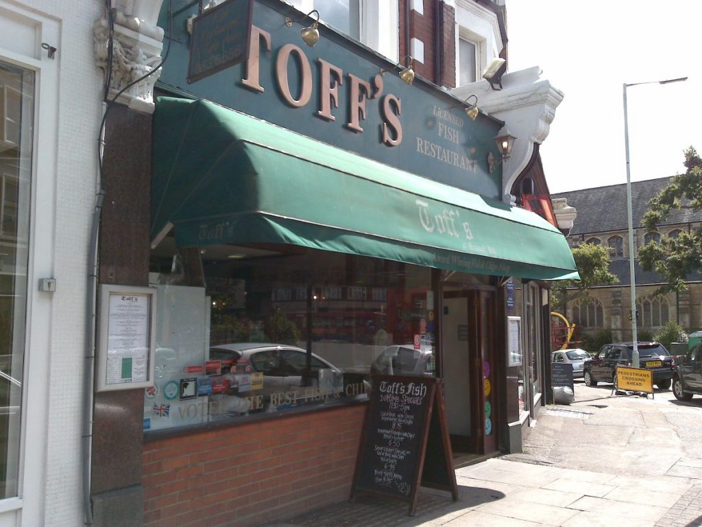 People walk past Toff’s Fish Restaurant in London, featuring a green awning, glass windows, and a chalkboard menu at the entrance.