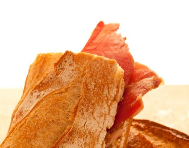 A crusty bread sandwich with red cured meat sits on a plain light surface,