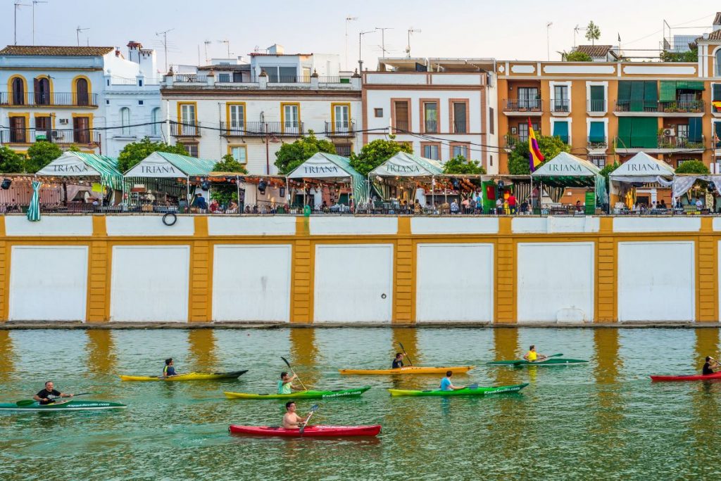 People kayak along the Guadalquivir River in Triana, Seville, passing vibrant buildings and outdoor cafes which are worth visiting in for the views and ambience