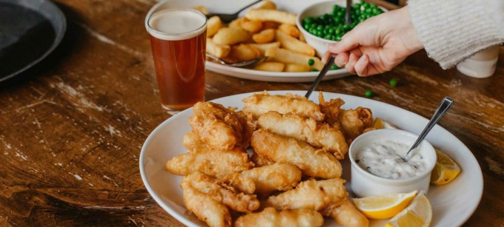 A hand reaches for battered fried fish with lemon wedges and tartar sauce at a London pub, surrounded by beer, fries, and peas.