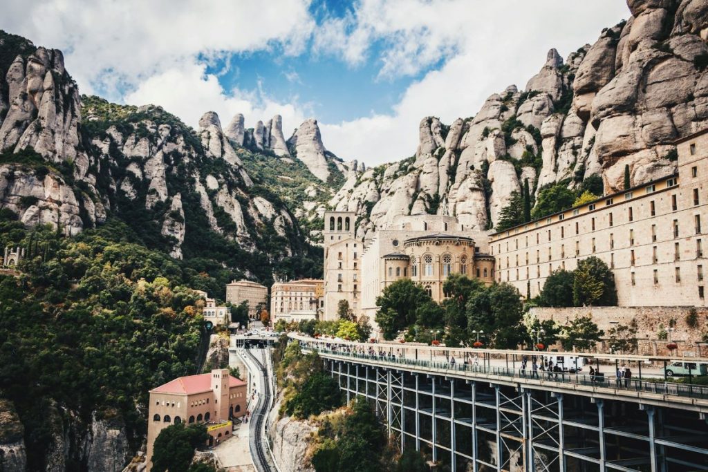 The Montserrat Monastery stands against jagged mountains in Spain, surrounded by green trees. Visitors walk along a pathway beneath blue skies.