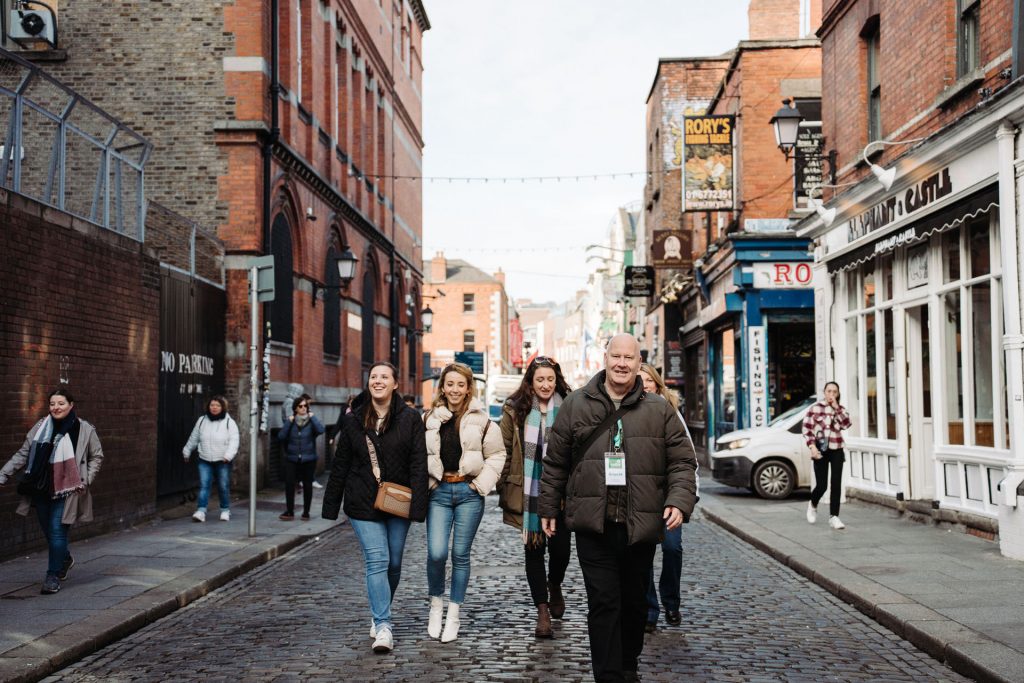 A group of people in winter coats walk along a cobblestone street lined with brick buildings and shops in Dublin.
