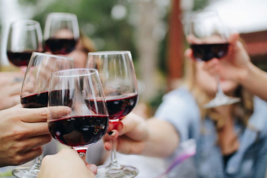 Several friends toast with glasses of red wine at a bustling outdoor wine bar in Milan, surrounded by greenery and laughter.