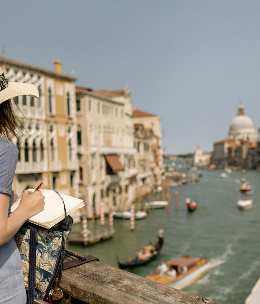 A woman in a sunhat writes in her notebook on a Venetian bridge, overlooking gondolas and the domed Santa Maria della Salute.