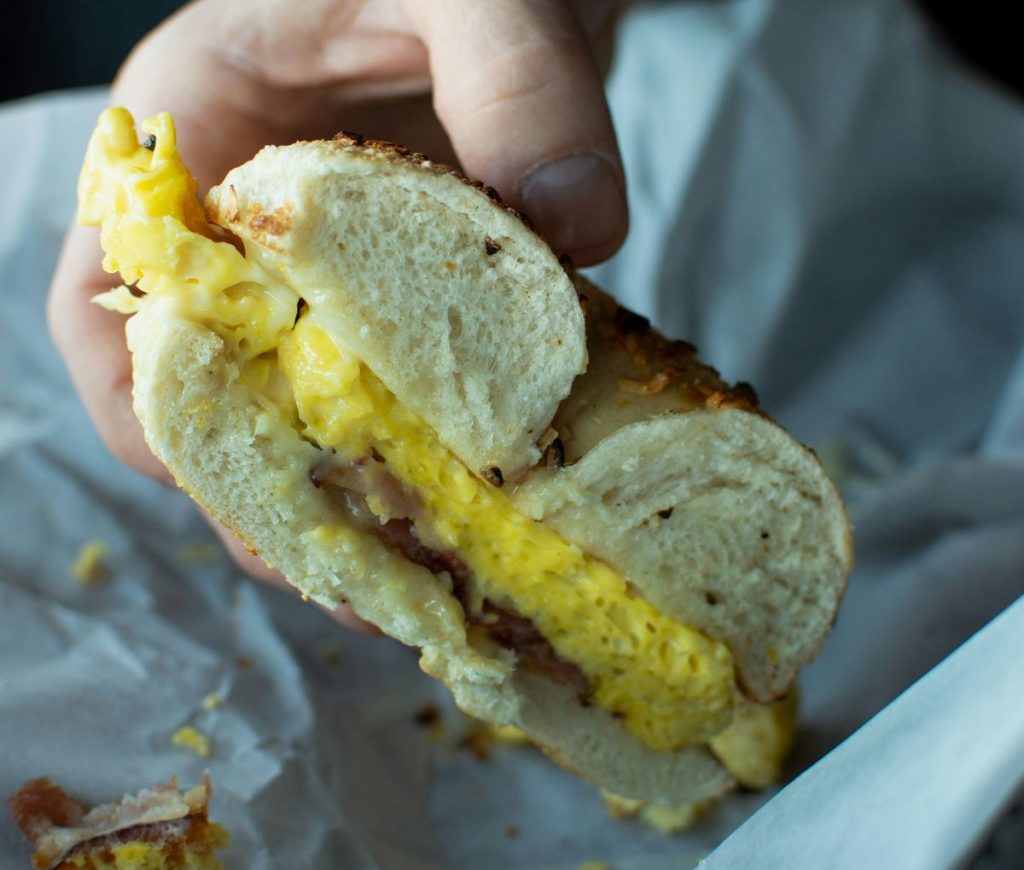 A person holds a breakfast sandwich with scrambled eggs, cheese, and bacon on a sesame seed bagel in London.