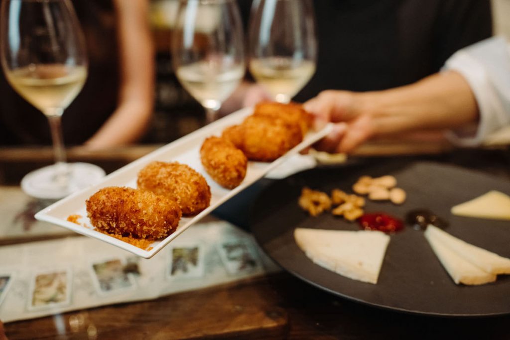 A hand serves golden, breaded croquettes onto a table with wine glasses, cheeses, nuts, and spreads near Madrid’s Prado Museum.