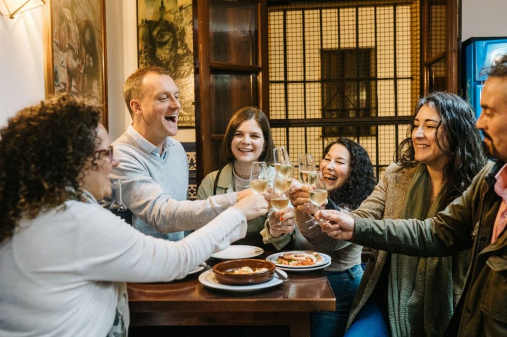 Six friends toast with drinks at a warmly lit restaurant, sharing smiles, laughter, and plates of food during dinner.