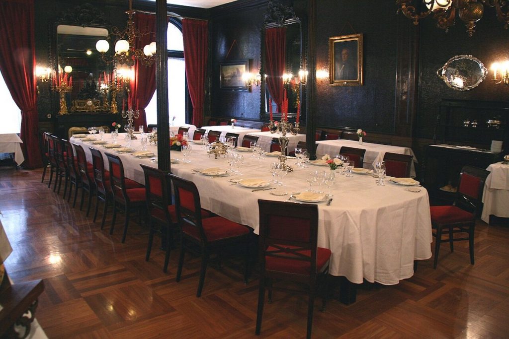 A formal dining room near the Prado Museum features red chairs, chandeliers, white tablecloths, large mirrors, and ornate dark walls.