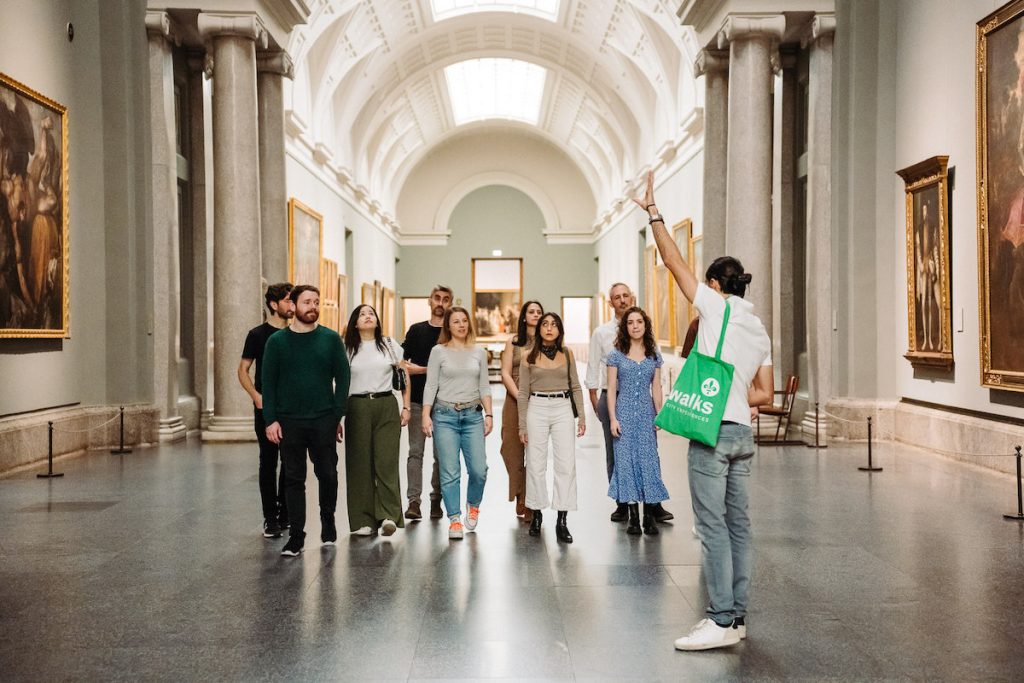 A tour guide with a green tote bag leads visitors through Madrid’s Prado Museum, discussing nearby restaurants amid classical paintings and marble columns.